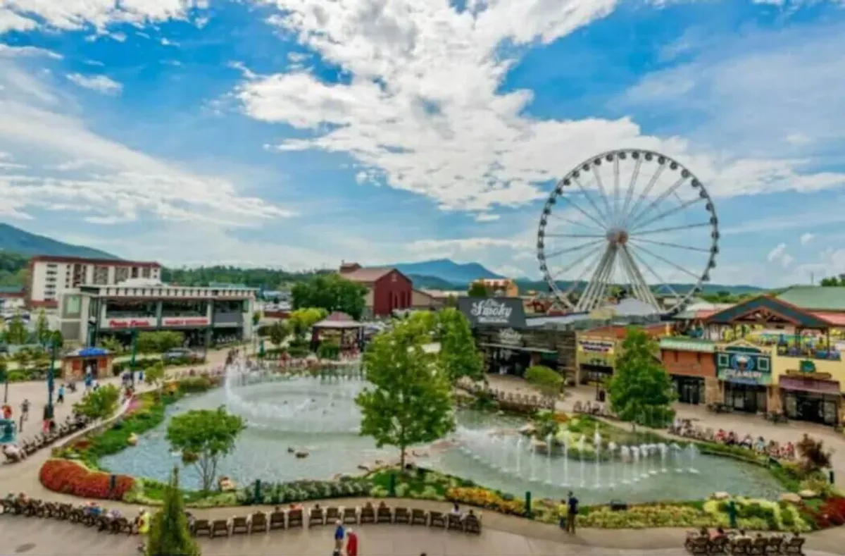 A bustling plaza is captured with a sparkling fountain, vibrant ferris wheel, pond, and nearby shops and restaurants. The scene is joyful and festive, populated by people biking, walking, and dining outdoors, evoking the spirit of shared group adventures.