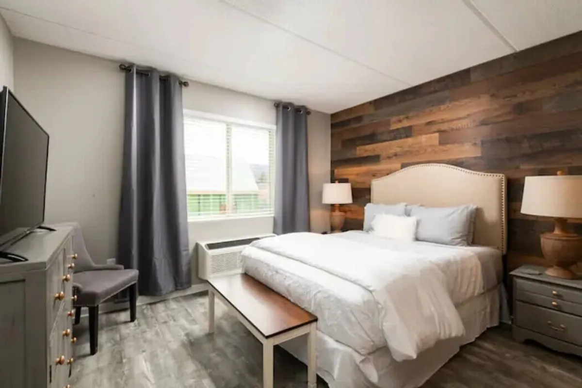 The image showcases a serene bedroom with a plush bed against a warm wood accent wall, soft sunlight streaming in through a large window, nightstands on either side, and a television perfectly positioned for cozy evenings. The space exudes comfort and tranquility, inviting guests to unwind or start their day in peaceful surroundings.