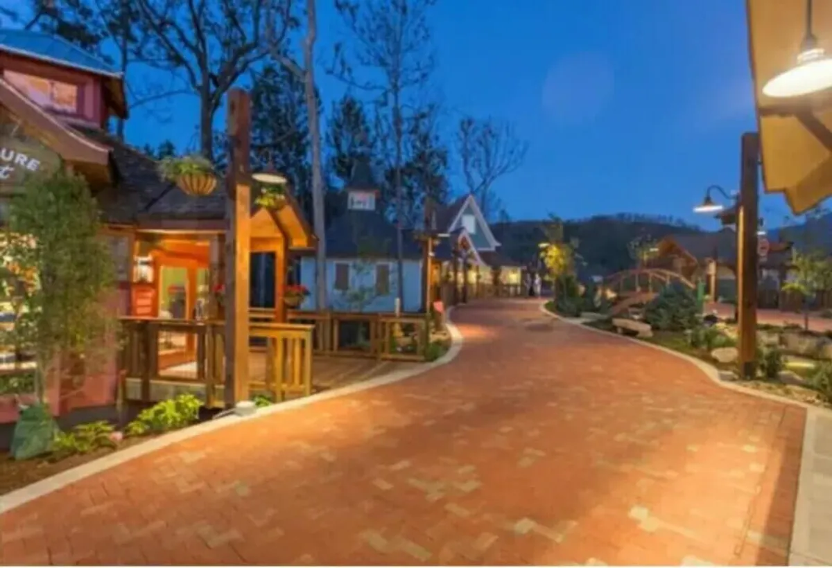 A charming cobblestone pathway winds between vibrant storefronts, lush greenery, and whimsical decorative lighting. A quaint bridge and lush planters invite leisurely strolls, setting a cozy, romantic scene perfect for couples exploring downtown.