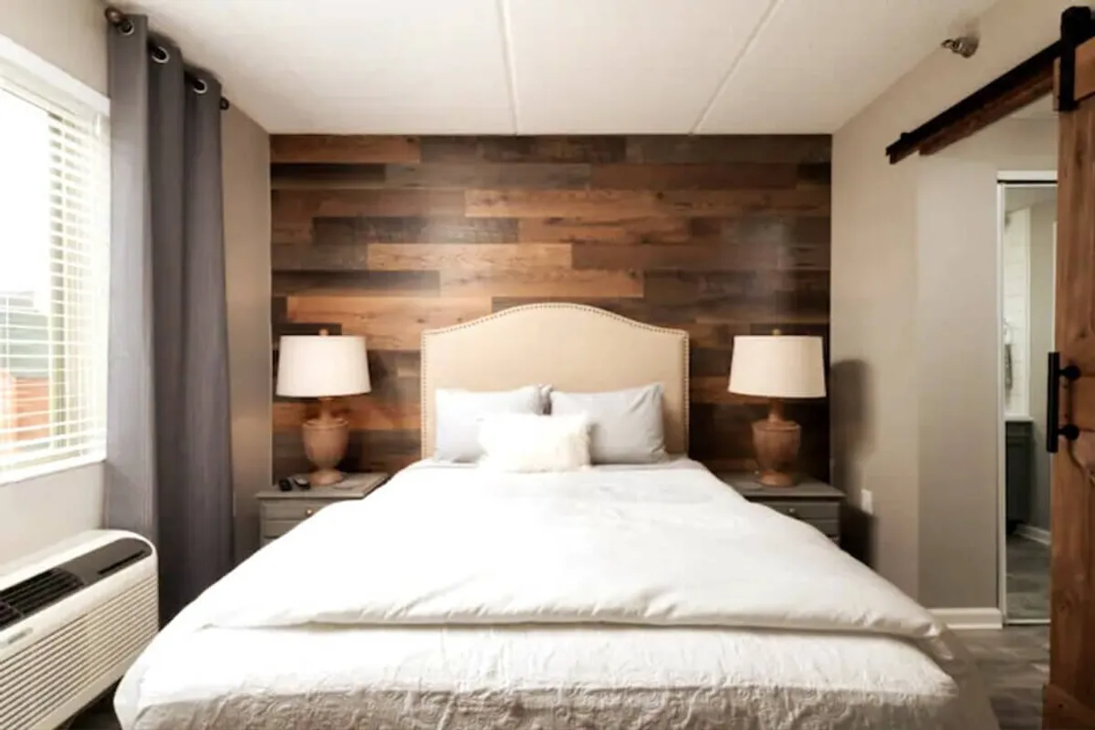 This image features a well-appointed bedroom with a bed dressed in plush, inviting bedding, a wooden accent wall, and softly glowing lamps. The scene radiates tranquility, emphasizing natural light and a restful atmosphere.