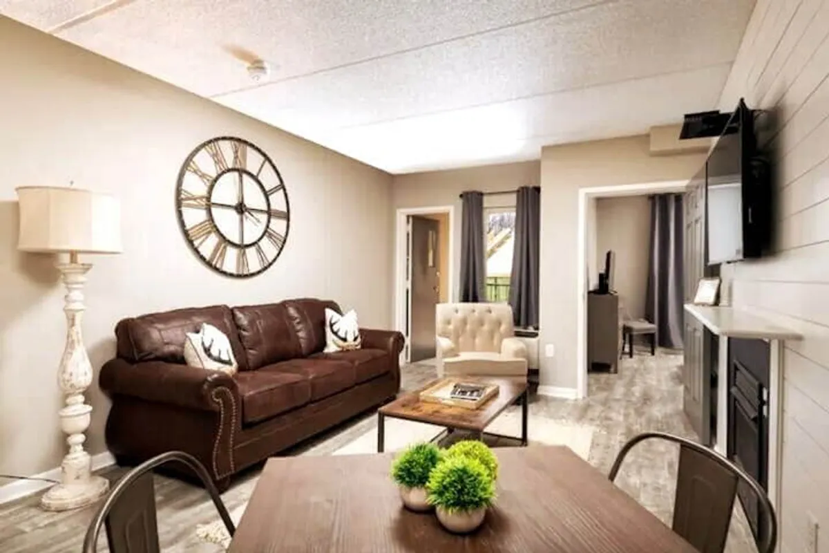 A cozy living area with a brown leather sofa, large wall clock, wooden coffee table, and lush green plants. The space is bathed in natural light, creating a warm and inviting environment ideal for entertaining guests, reading, or enjoying a quiet break.