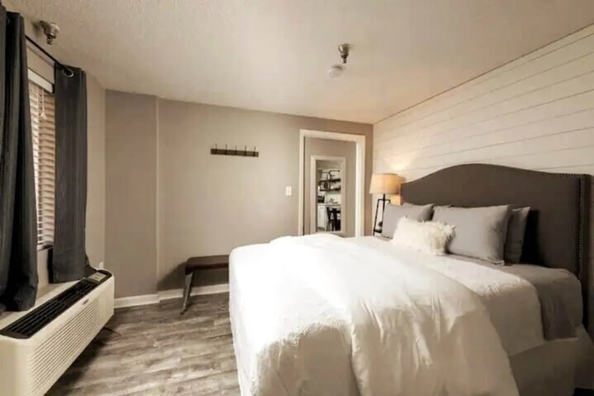 This image presents a king-sized bed with soft linens, a lampshade, and a window dressed with room-darkening curtains. The overall mood is restful and cozy, ideal for a good night's sleep.