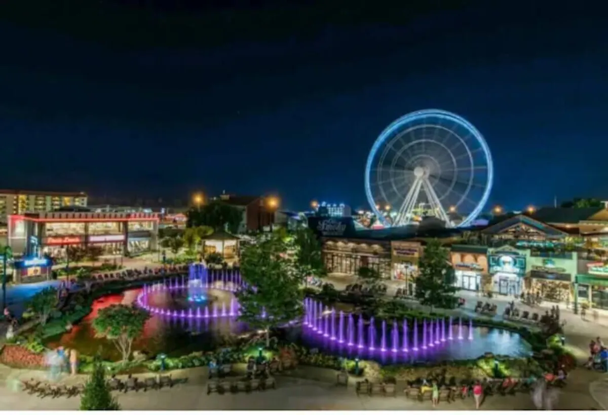 A lively Ferris wheel stands tall beside dancing colorful fountains, bustling restaurants, and inviting shops. The area is alive with energy, perfect for evening adventures and shared delights.