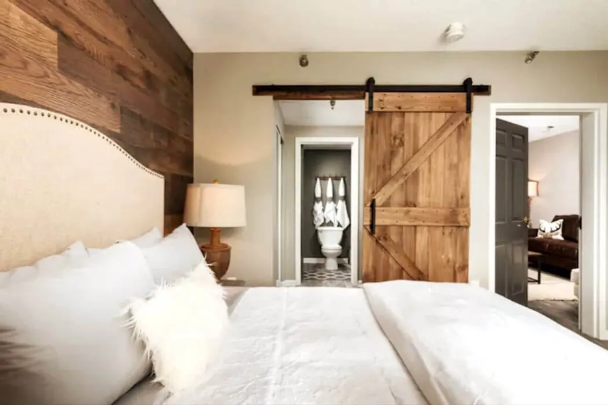 This bedroom showcases a comfortable bed loaded with plush pillows, a rustic wooden barn door, and a gentle table lamp. The lighting and decor evoke calm, inviting guests to unwind with a book or simply relax.
