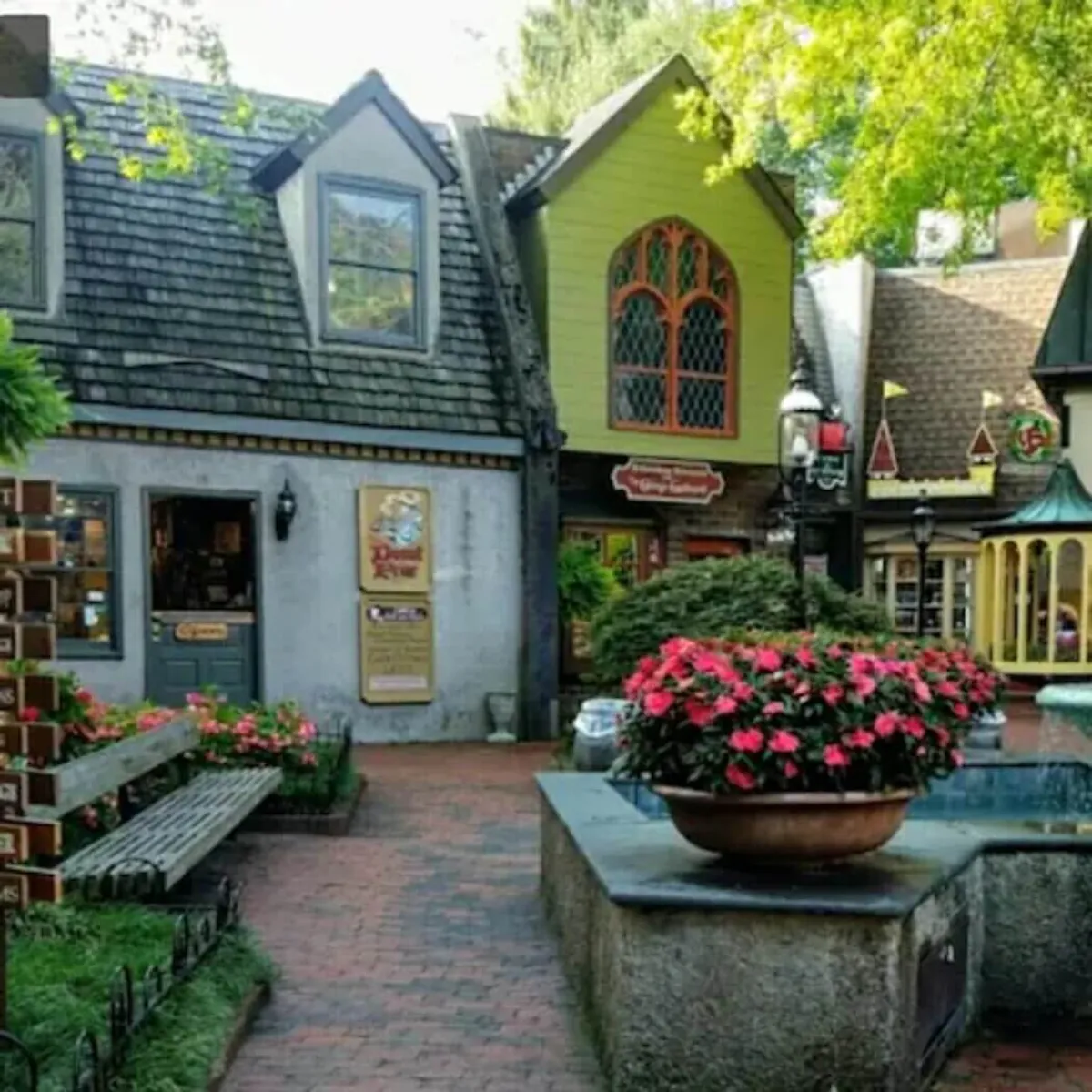 A cheerful street scene features colorful storefronts, abundant flower planters, and a water fountain. People are seen relaxing and socializing, highlighting the area's approachable and welcoming vibe. The setting suggests a perfect backdrop for daytime group adventures, shopping trips, and casual meetups.