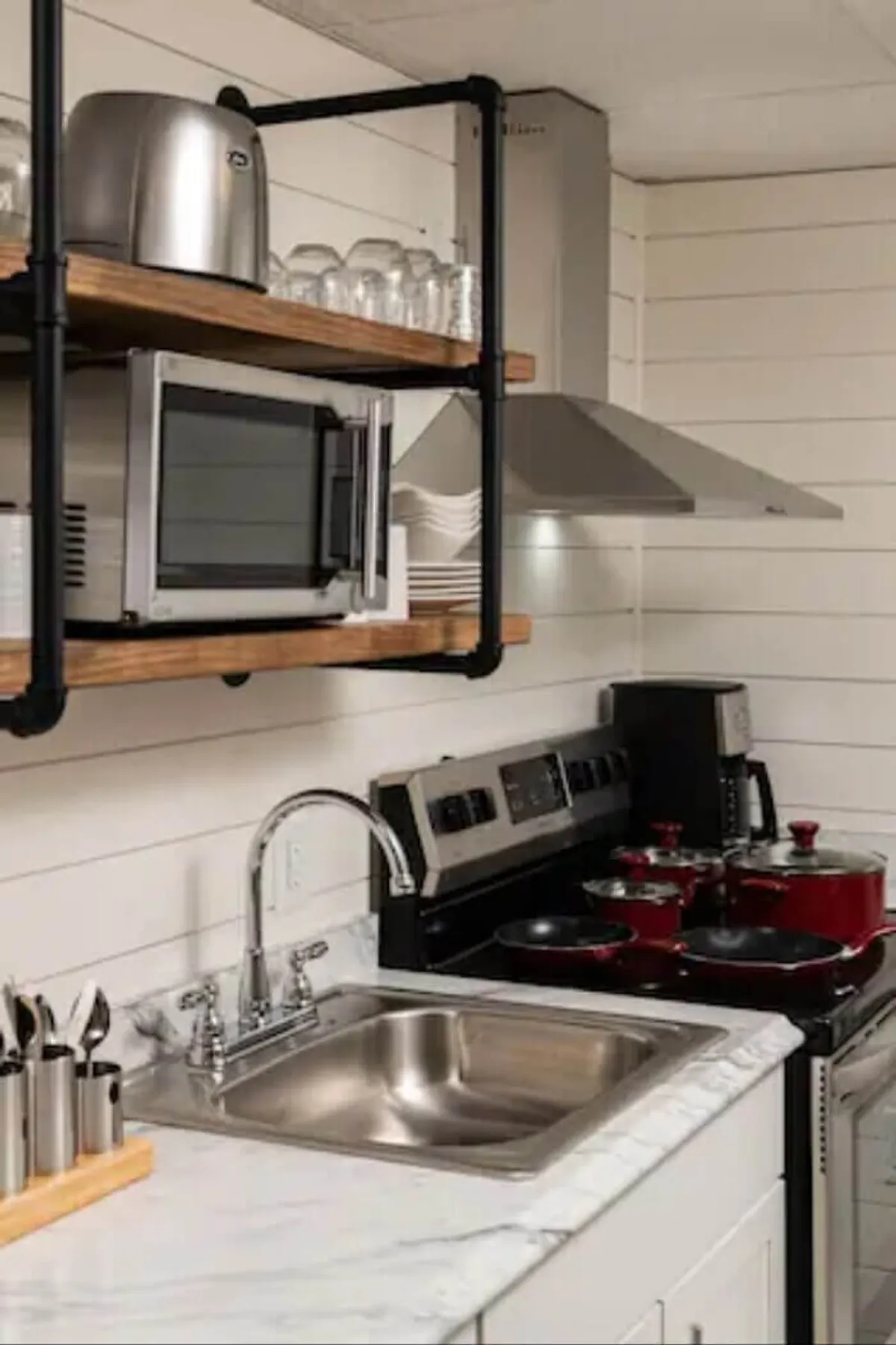 An inviting kitchen area with a stainless steel sink, red cookware, and open shelving filled with glassware. The space appears bright and conducive to both daily routines and special occasions, highlighting the kitchen’s open connection to communal living areas.