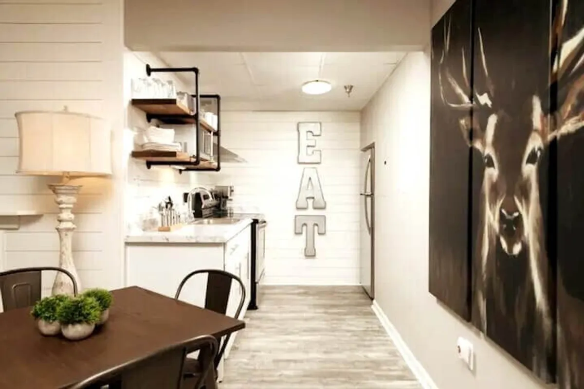 The image highlights a dining table set within an inviting kitchen area, with deer artwork and 'EAT' wall decor adding personality. The setup suggests a space for shared meals and socializing, tying directly into family gatherings around the table.
